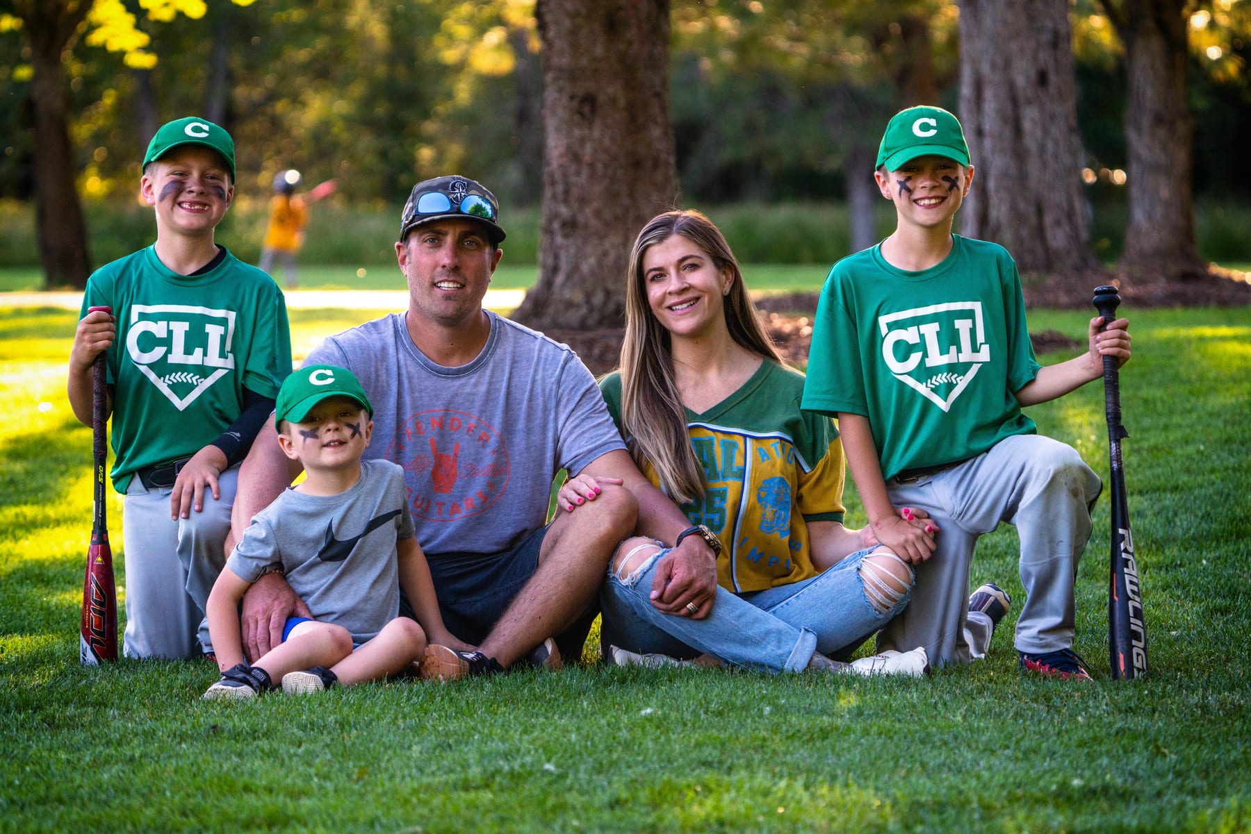 A family in matching ball caps sits together on the grass with bats and gloves.