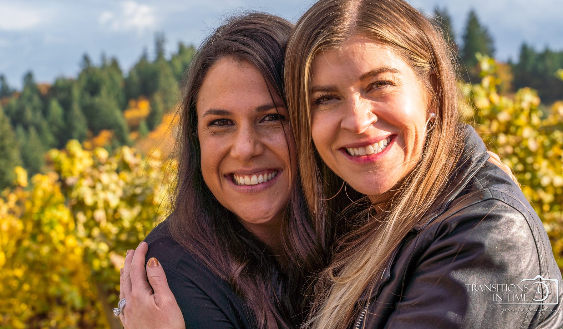 A mother and daughter lean their heads together among autumn vines.