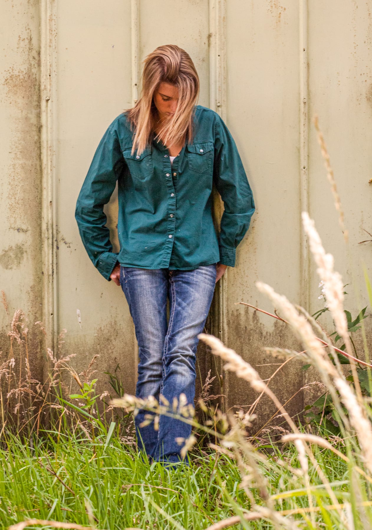 A senior leans against a weathered barn wall in tall grass.