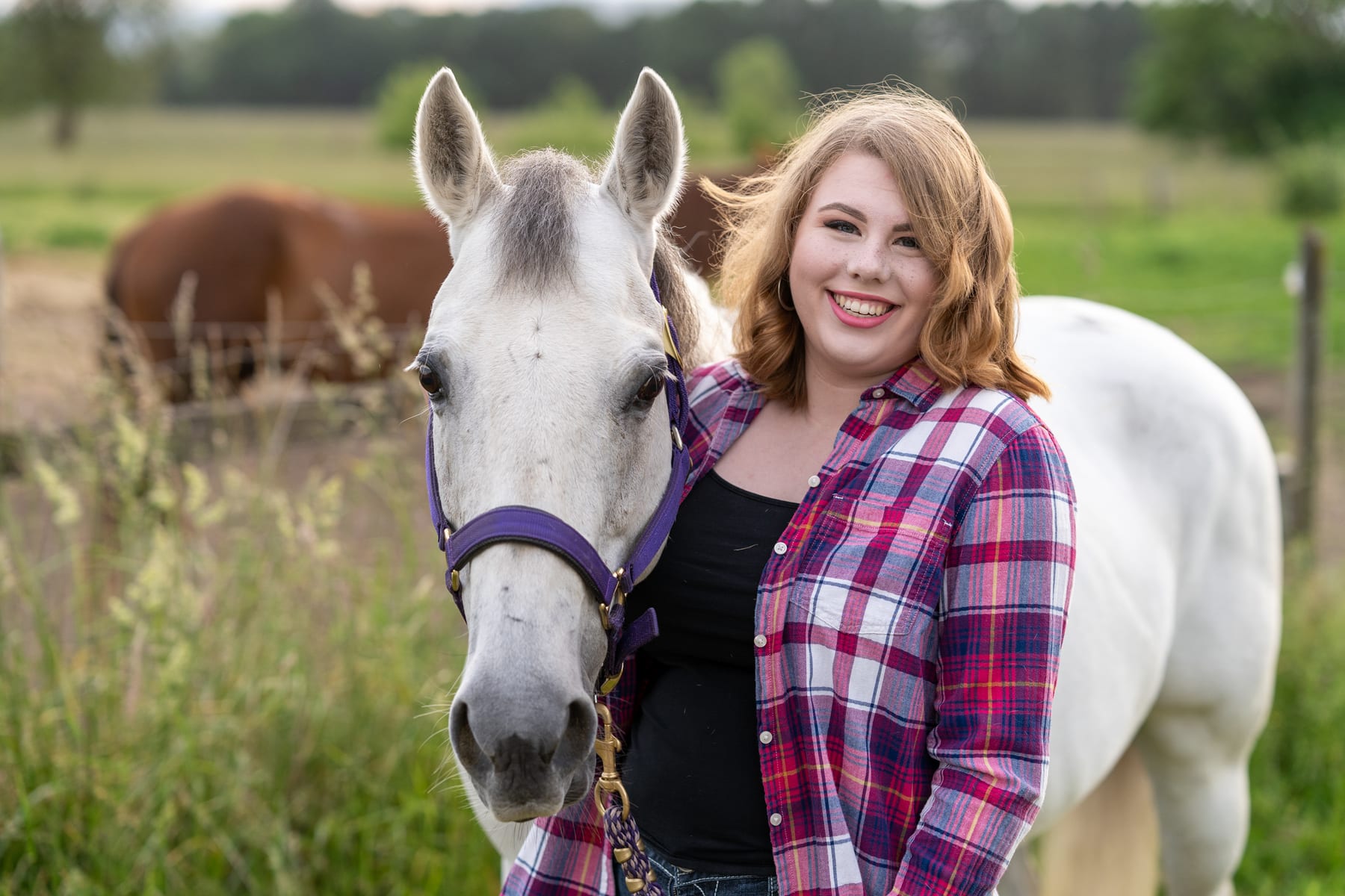 A senior in a plaid shirt stands with her horse in a green field.