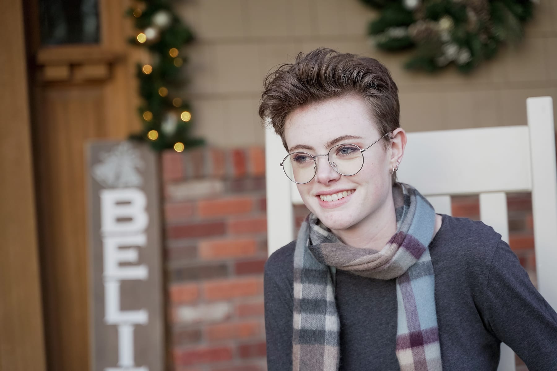 A senior in glasses and a scarf smiles on a porch decorated for the holidays.