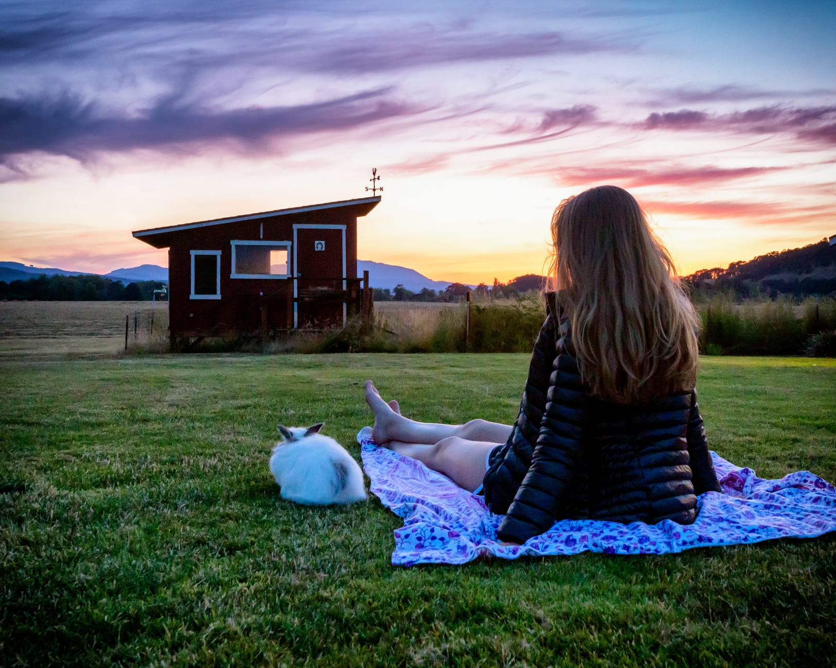 A senior sits on a blanket at sunset looking toward a small cabin.