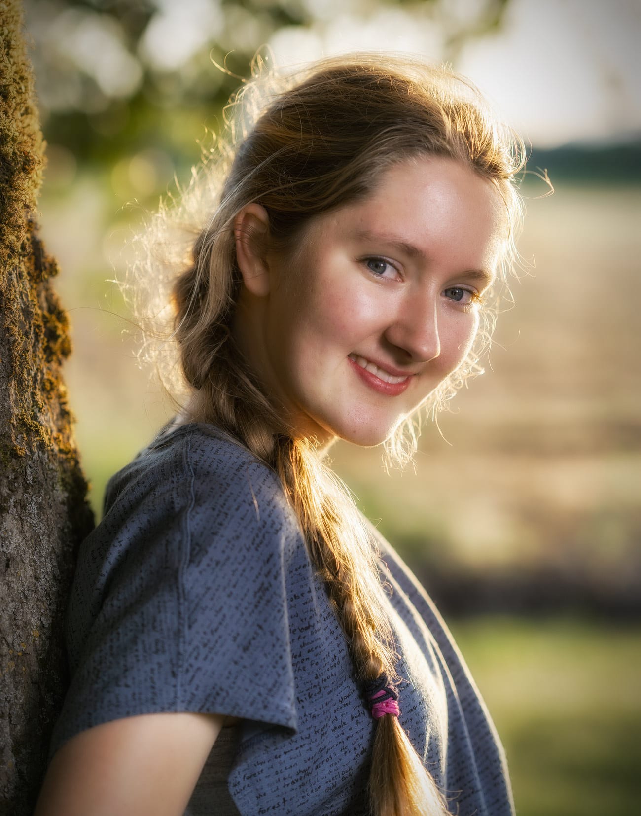 A senior smiles over her shoulder beside a tree in soft light.