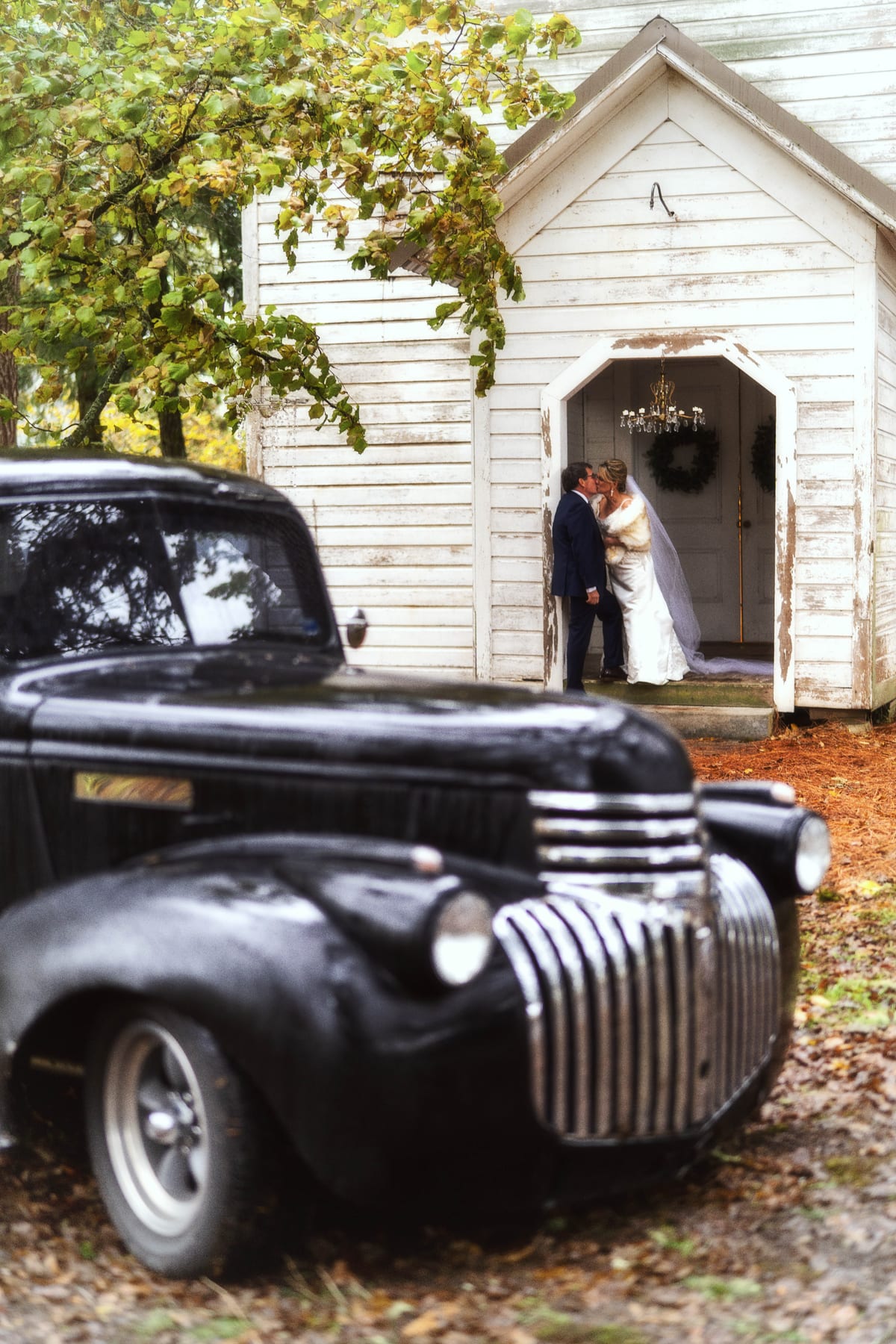 A couple stands at the door of a white country chapel beside a vintage black truck.