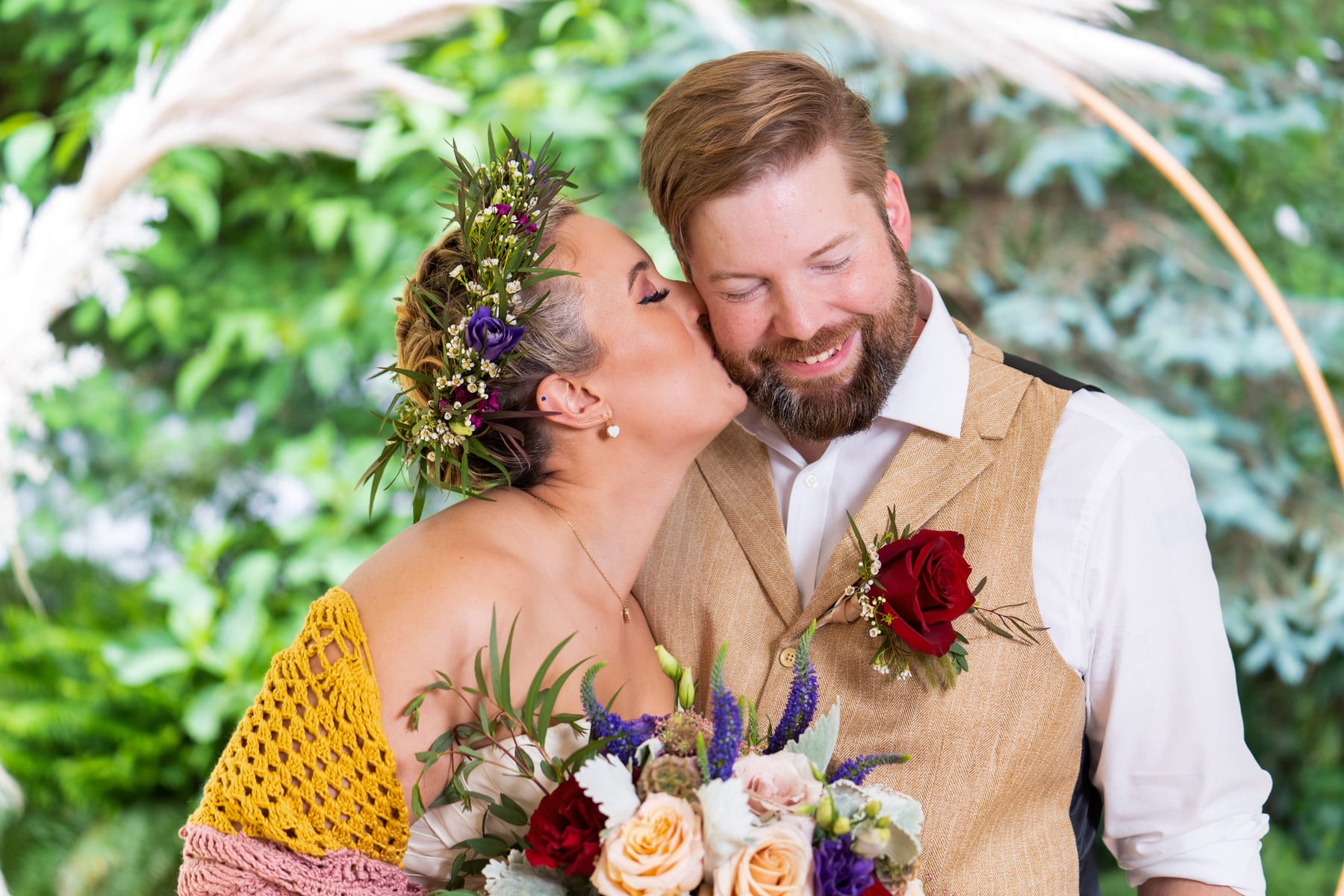 A groom kisses his bride on the forehead as she holds a wildflower bouquet.
