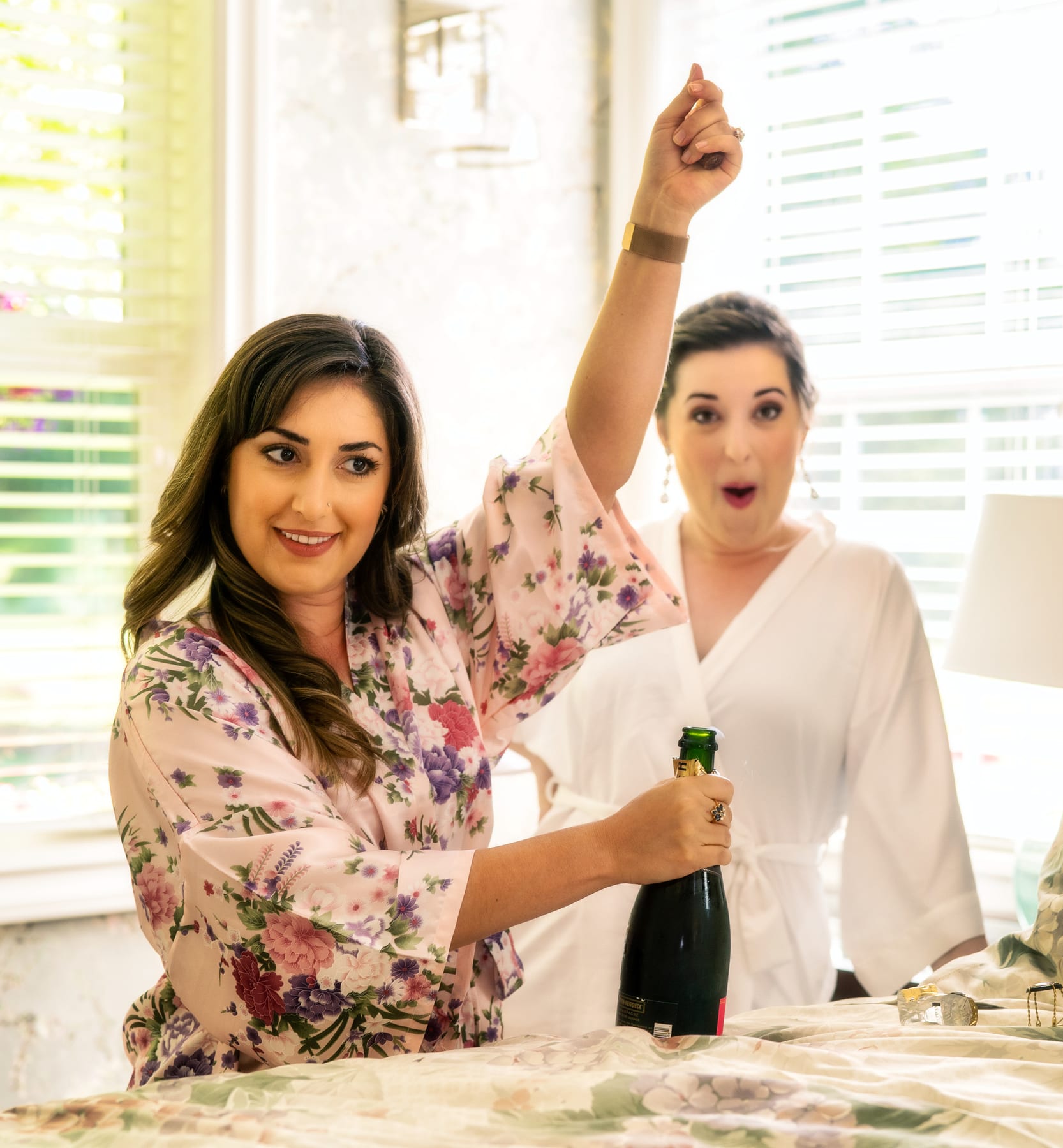 A bride and her friend cheer while opening champagne as they get ready.