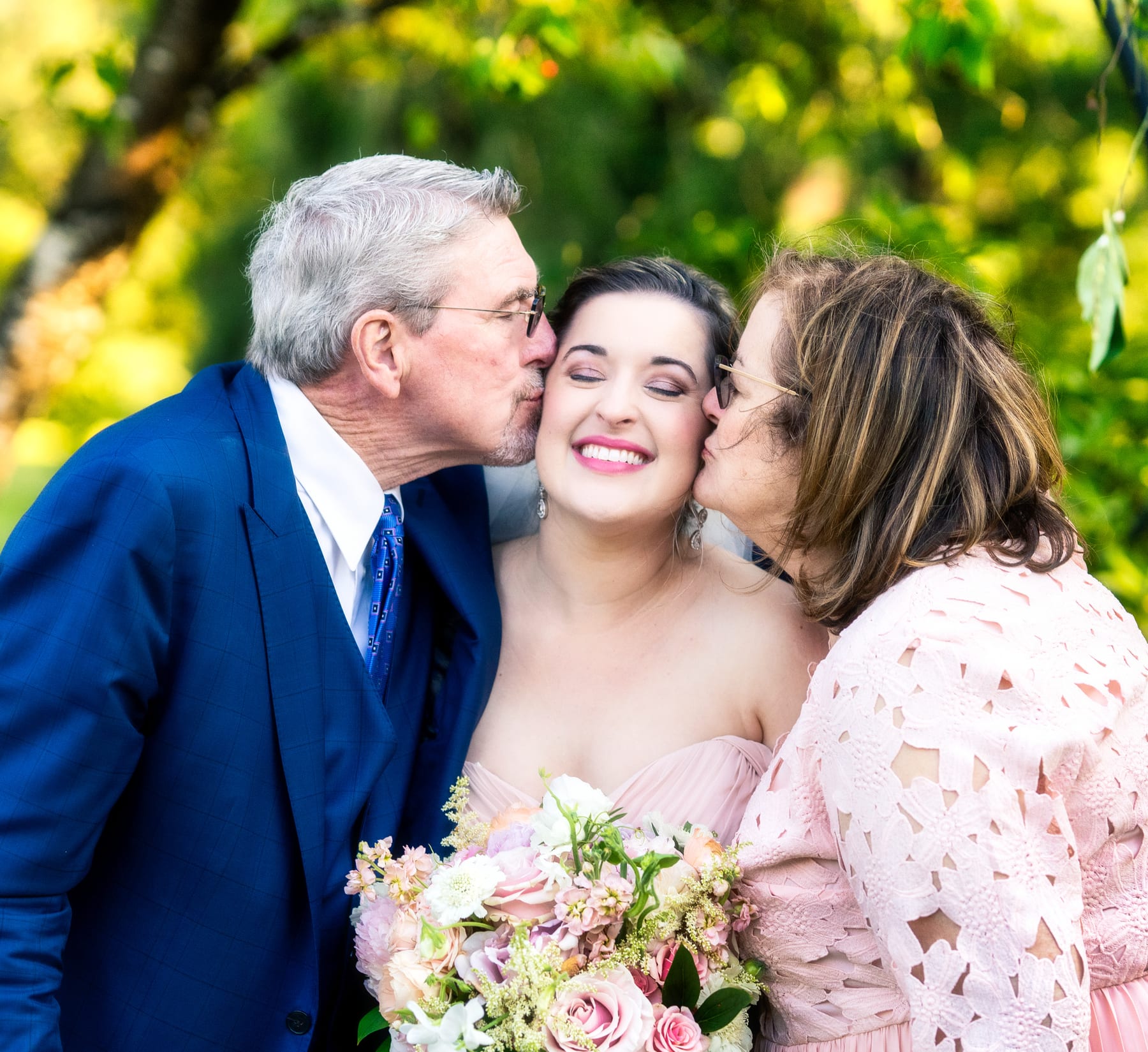 Two parents kiss their daughter's cheeks on her wedding day while she laughs.
