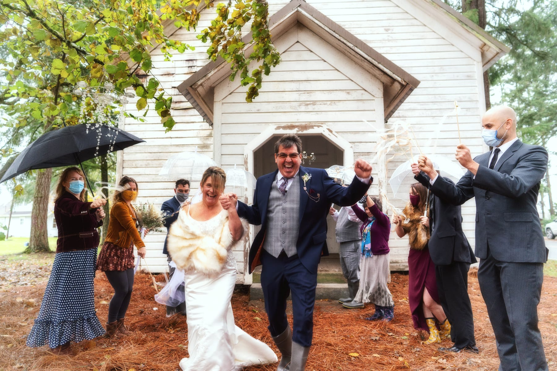 Guests cheer under umbrellas as a couple leaves a country chapel in the rain.