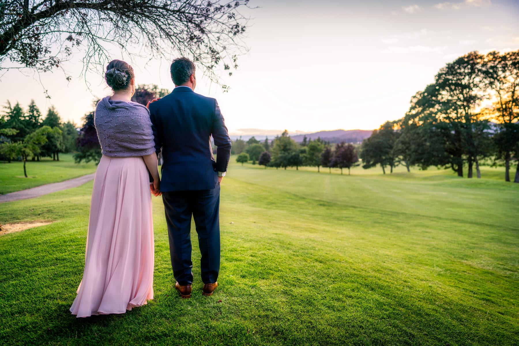 A newly married couple stands arm in arm looking out over a golf course at sunset.