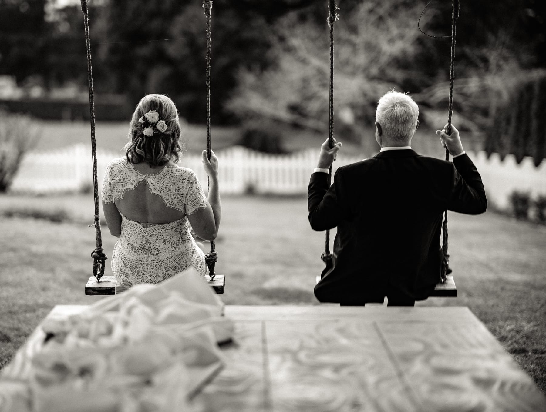A black and white photograph of a couple side by side on garden swings.