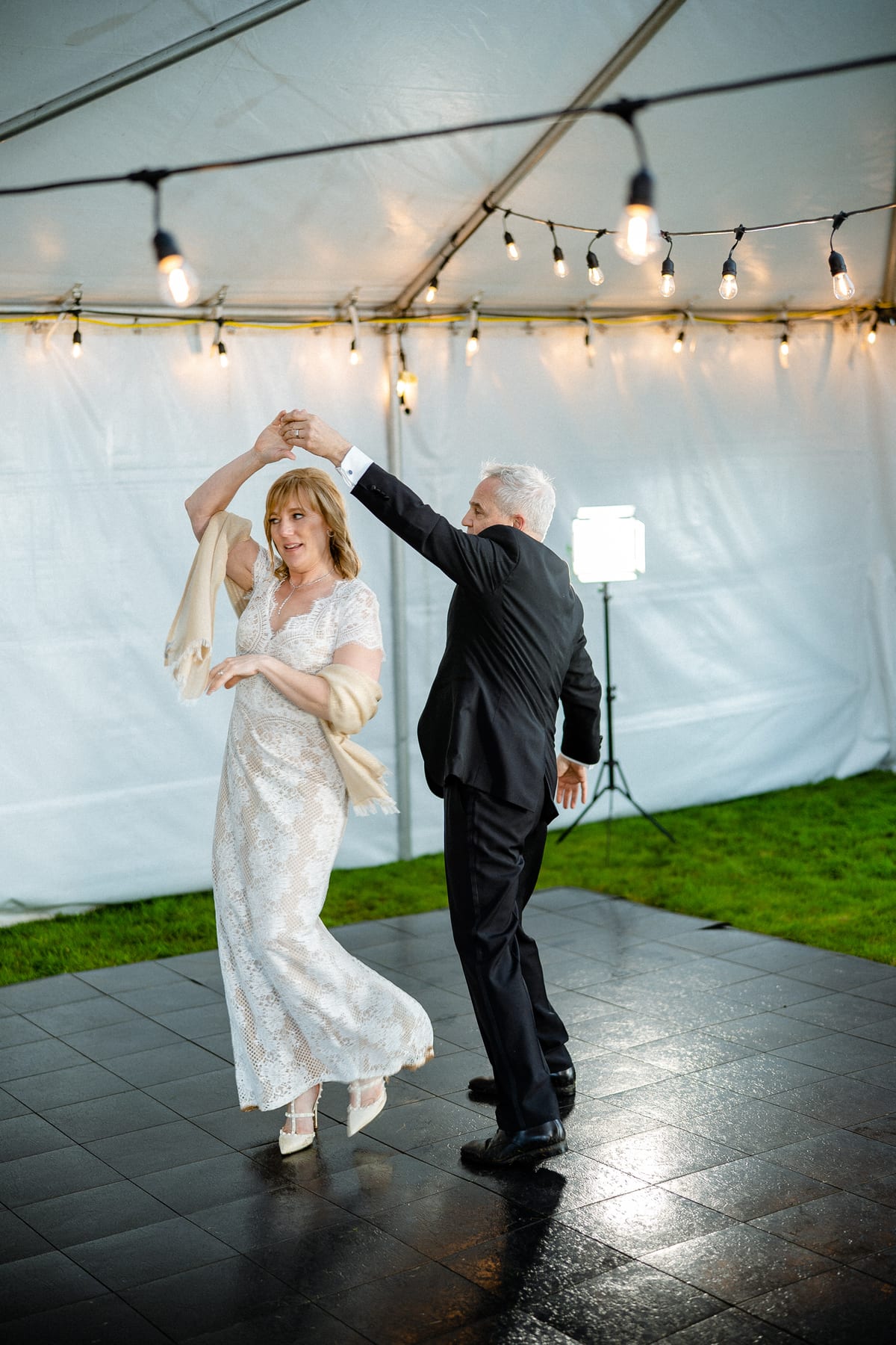 A couple dances beneath string lights in a reception tent.