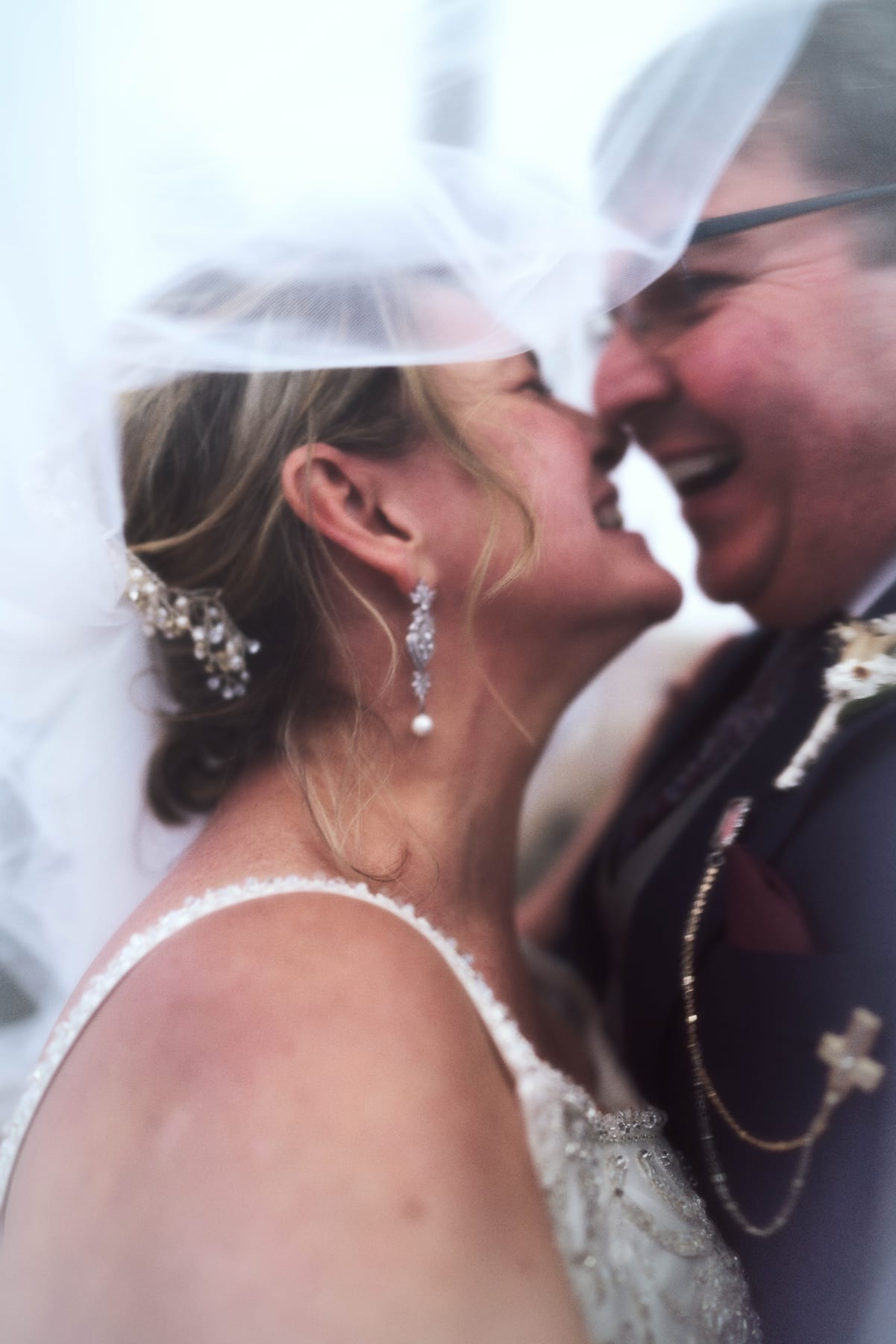 A couple touches foreheads and laughs beneath the wedding veil.