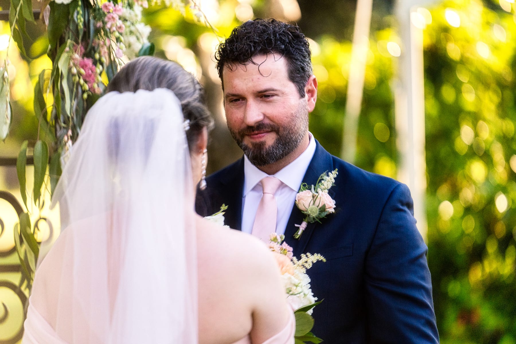 A groom looks at his bride during their vows beneath a floral arch.